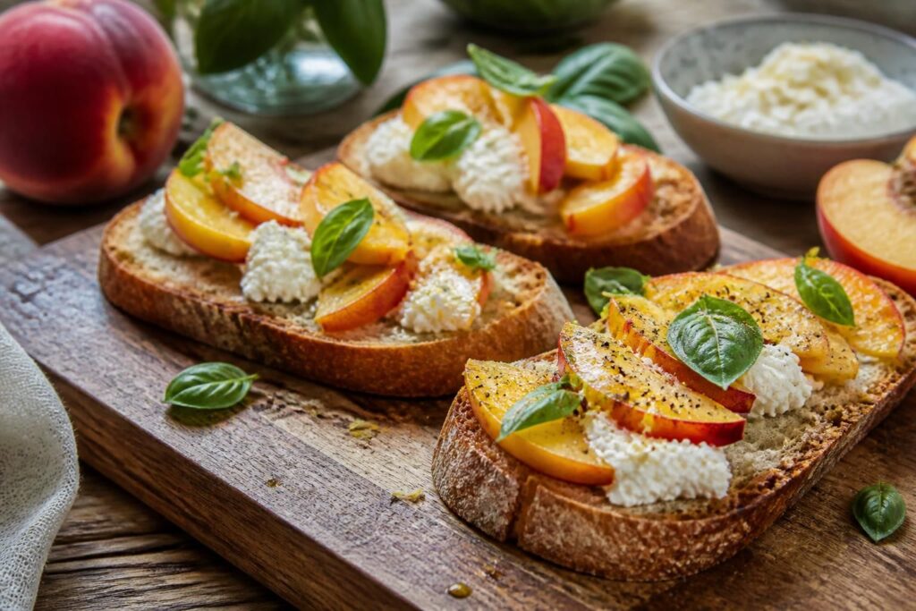 Bruschette con pesche fresche, ricotta cremosa e foglie di basilico su un tagliere di legno, aperitivo estivo dolce-salato